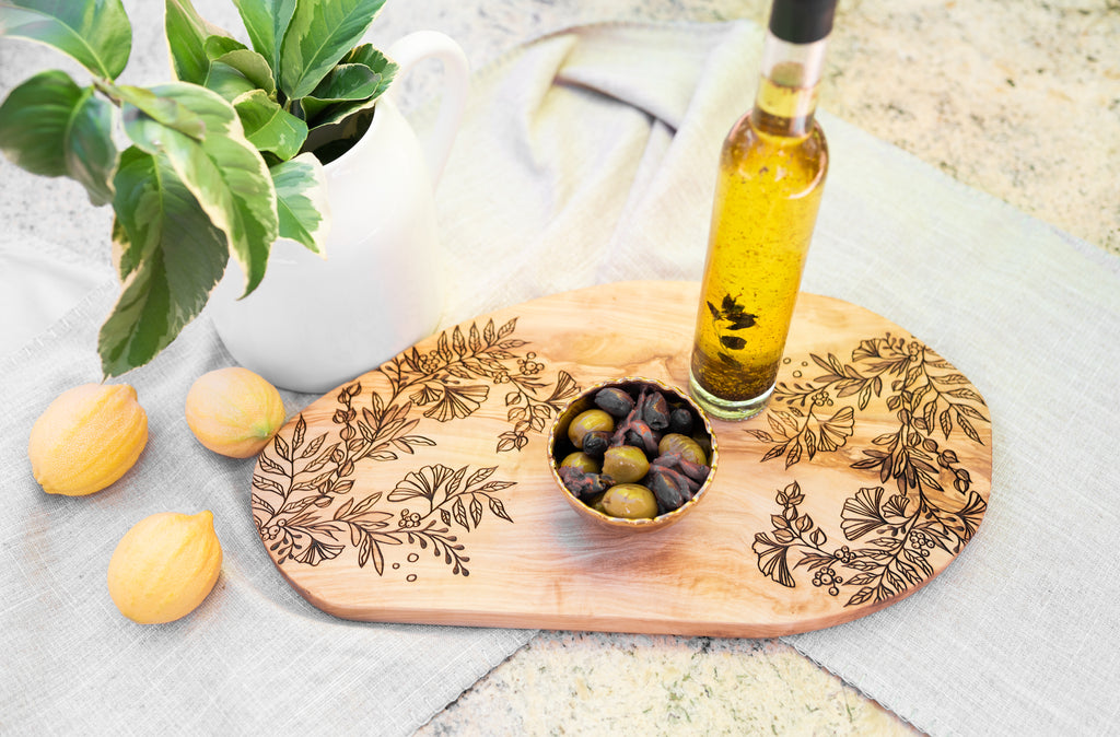 Wooden cutting board with olive design, bottle of olive oil, and olives on a light surface.