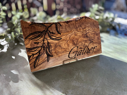 Live-edge olive wood serving board with olive leaves engraving angled in the sun
