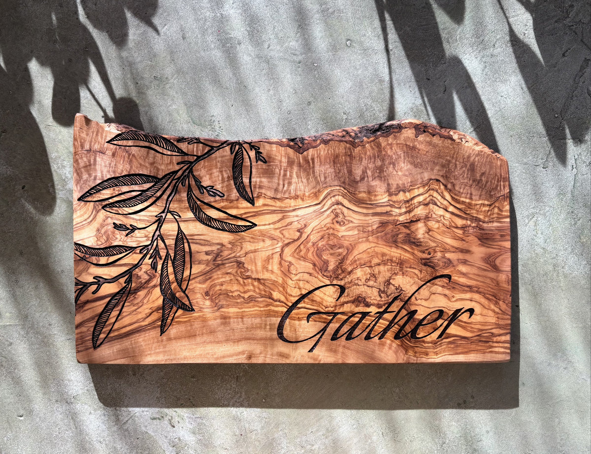 Wooden cutting board with engraved leaves and 'Gather' text on a textured surface