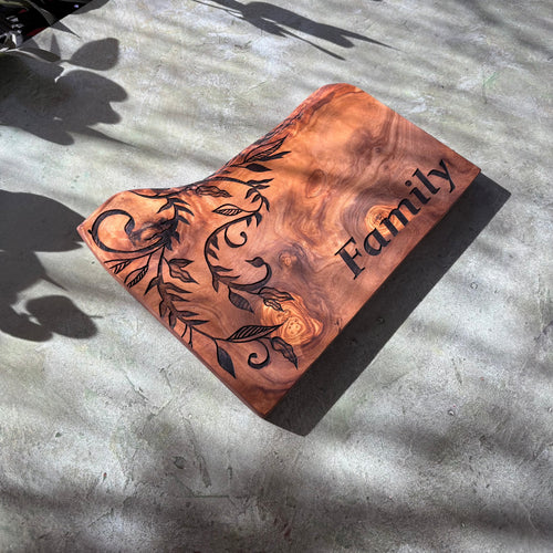 Angled view of a rustic olive wood serving board engraved with Family and a botanical design